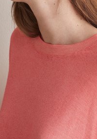 Zbliżenie osoby w koralowym, dzianinowym swetrze z okrągłym dekoltem, z jasnobrązowymi włosami widocznymi po lewej stronie.