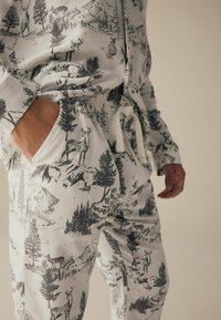 Das Loungewear-Set besteht aus einem weißen Stoff mit schwarzen Illustrationen von Wildtieren und Bergen, einem elastischen Bund und einer Gürtelschlaufe, die die filigranen Muster hervorhebt.