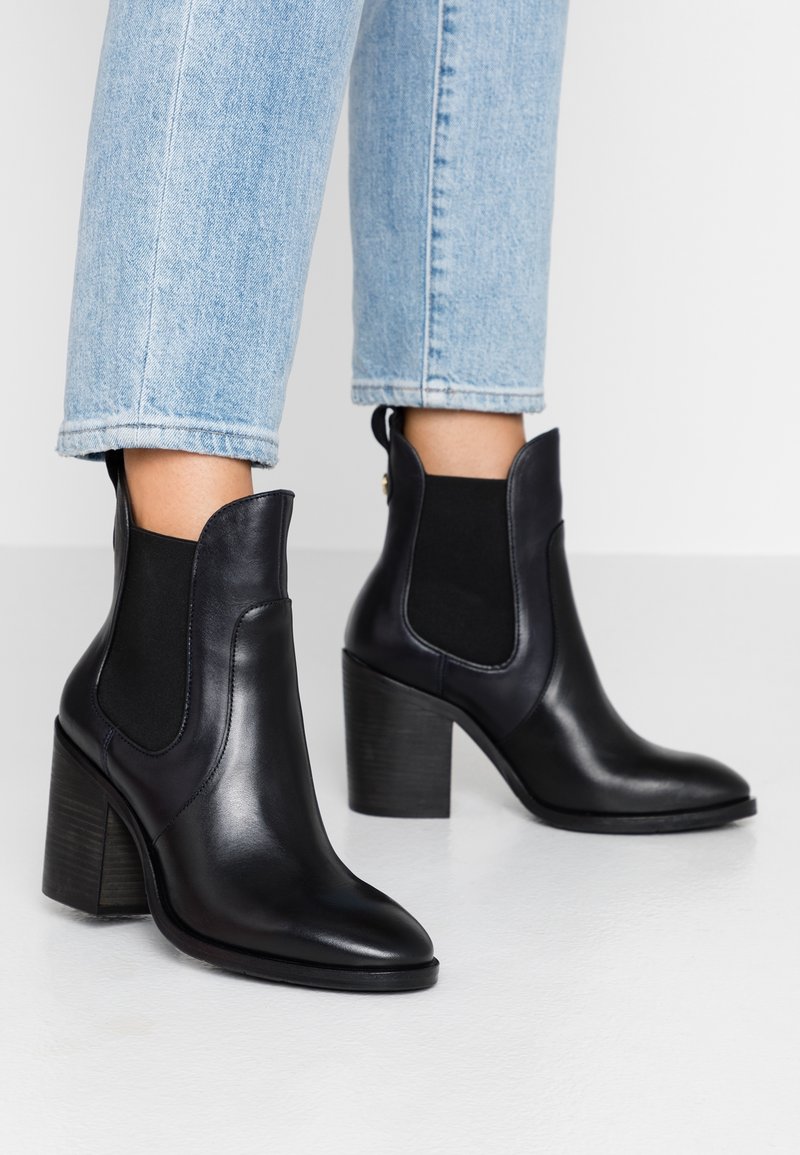 Bottes basses en cuir noir avec des panneaux élastiques sur les côtés, un bout pointu, et un talon épais texturé. Portées avec un jean bleu clair retroussé.