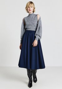 Grauer Strickpullover mit Ausschnitt an der Schulter, kombiniert mit einem marineblauen A-Linien-Midirock und schwarzen kniehohen Stiefeln mit glattem Finish.