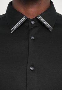 Polo shirt nera con una texture liscia, caratterizzata da un colletto nero con logo bianco "Emporio Armani" e bottoni neri sulla parte anteriore.