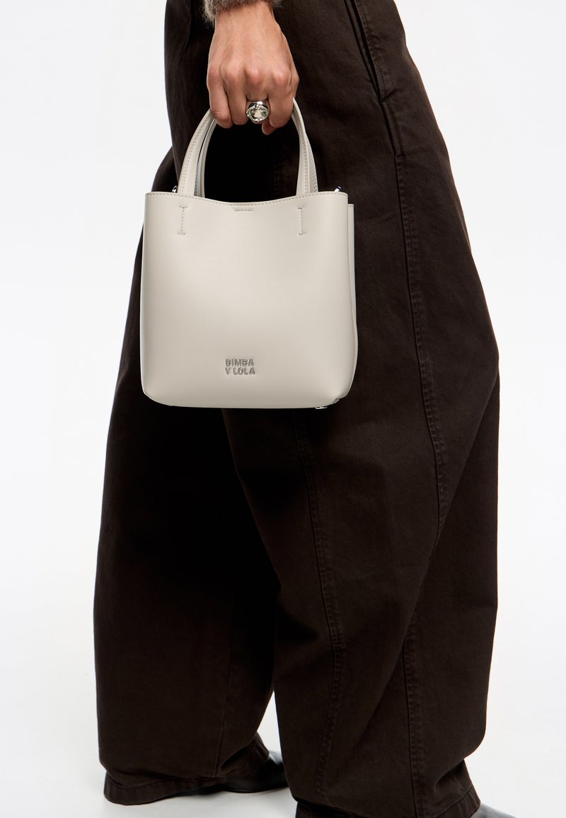 Bolso de mano de cuero gris claro con un diseño estructurado, que presenta dos asas cortas, un cierre de cremallera y un logo en relieve en la parte inferior.