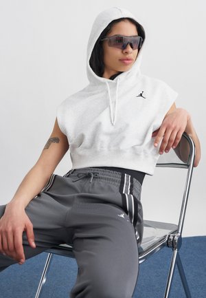 Młoda osoba ubrana w szarą bezrękawnikową bluzę z kapturem z logo Jordan, ciemne sportowe spodnie, okulary przeciwsłoneczne, siedząca zrelaksowana na metalowym krześle na szarym tle.