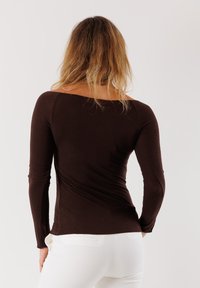 Top lungo marrone con maniche lunghe e scollatura a barca, design aderente e tessuto elasticizzato; abbinato a pantaloni bianchi, mostrando una texture liscia.