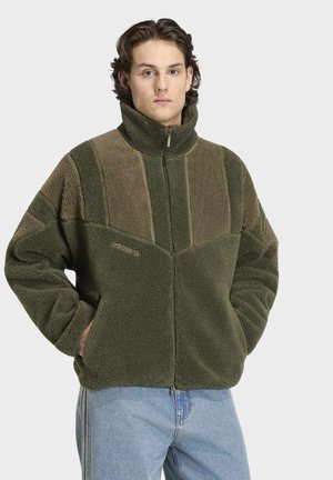 Jeune homme aux cheveux bruns portant une veste polaire verte et marron avec un col montant et un jean bleu clair, les mains dans les poches.