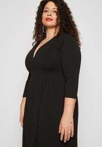 Vestido negro con escote en V profundo, mangas tres cuartos y detalles de pliegues en la cintura. Tejido suave y elástico con una silueta elegante.