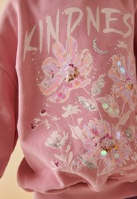 Rosa sweatshirt med blommig paljettdesign i pastellfärger, med ordet "KINDNESS" framträdande i en ljus font.