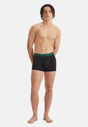 Junger Mann mit Sommersprossen und kurzen Dreadlocks, der schwarze Boxershorts mit grünem Bund trägt, barfuß vor weißem Hintergrund steht.