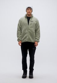 Ανοιχτοπράσινο φλις μπουφάν με φερμουάρ μπροστά και τσέπη, φορεμένο πάνω από μαύρο πουκάμισο και παντελόνι, σε συνδυασμό με γκρι σκουφάκι και σκούρα μποτάκια.