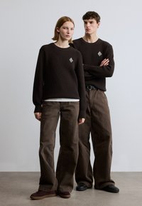 Pulls en maille côtelée marron avec des empiècements en forme de losange, associés à un pantalon marron à jambes larges. Les chaussures sont des baskets foncées.