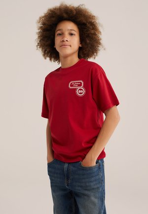 Jeune personne aux cheveux bouclés portant un t-shirt rouge avec deux logos et un jean bleu, debout les mains dans les poches devant un fond neutre.