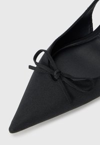 Scarpa piatta nera in tessuto con punta affusolata e un piccolo dettaglio a fiocco sulla parte anteriore. Interno liscio ed esterno texturizzato.
