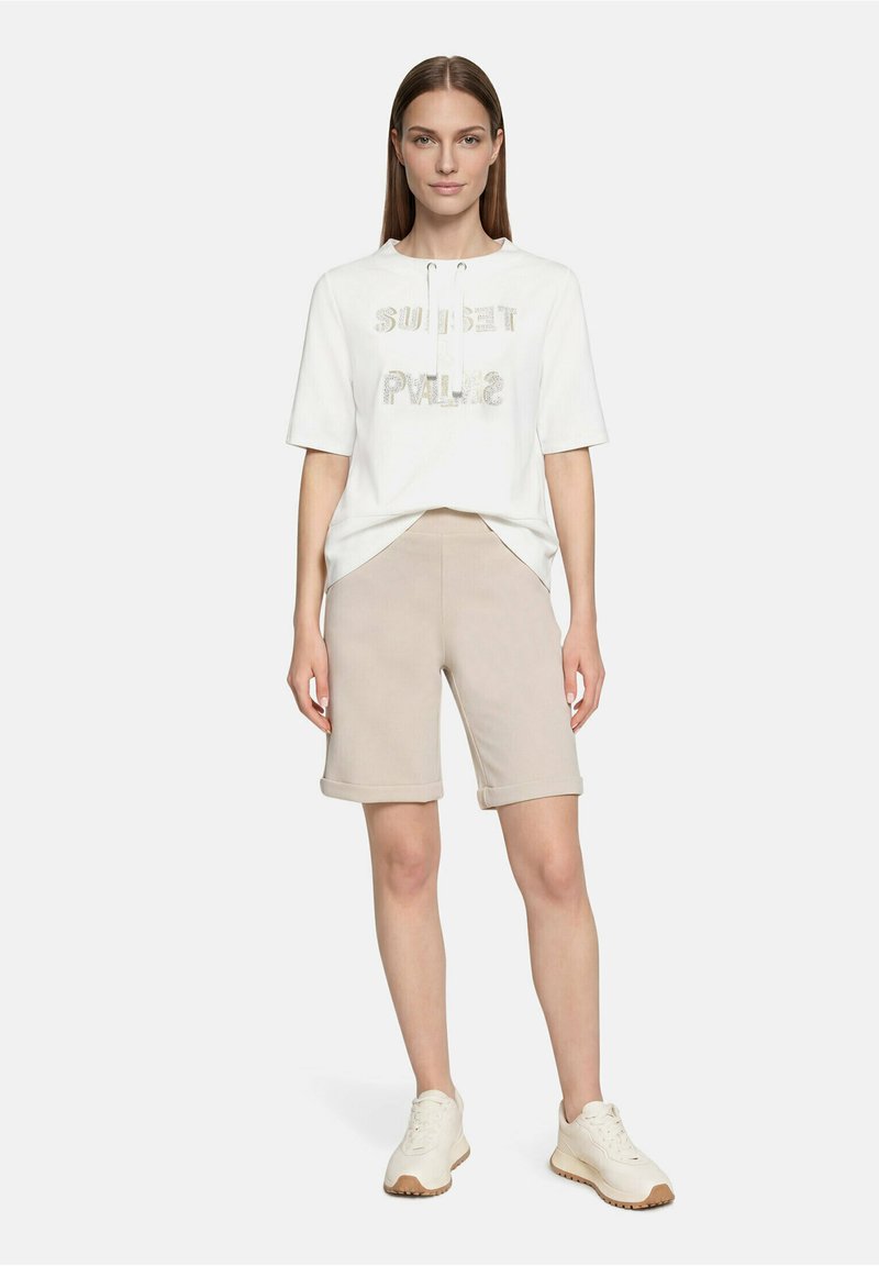 Femme portant un sweat à capuche blanc à manches courtes avec le texte « Sunset Palm », un short beige longueur genou et des baskets blanches, debout devant un fond uni.