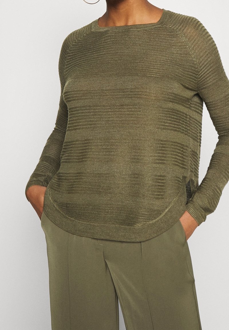 Personne portant un pull en maille côtelée vert olive à manches longues et un pantalon ample assorti, mains dans les poches.