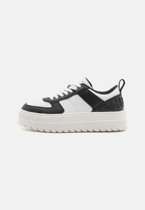 Zapatilla baja con paneles en blanco y negro, cordones blancos, suela gruesa blanca con estrías y lengüeta en el talón con letras en relieve.
