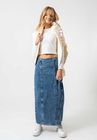 Denim maxi szoknya egyenes szabással, oldalsó gombokkal és világoskék mosással. Fehér croptop-pal és világos színű sportcipőkkel párosítva.
