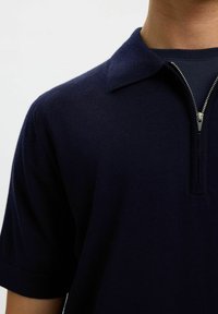 Gros plan sur une personne portant un polo sombre bleu marine à fermeture éclair, à manches courtes et col replié.