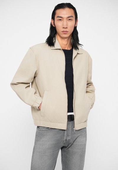 Άνδρας που φοράει μπεζ ζακέτα με φερμουάρ πάνω από μαύρο πουκάμισο και γκρι τζιν, στέκεται με τα χέρια στις τσέπες του μπροστά από έναν απλό φόντο.