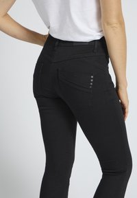 Jeans negri elastici, cu croială mulată, două buzunare în spate și nasturi decorativi pe buzunarul din stânga. Textură fină a materialului.