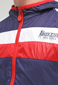 Námořnická, červená a bílá větrovka s vysokým límcem, předním zipem a logem na hrudi. Hladká textura s barevným blokovým designem a lesklým povrchem.
