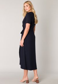 Marineblaues, kurzärmliges Midikleid mit einem Bindegürtel in der Taille. Weicher Stoff mit glatter Textur. Sandalen mit Absatz und klobigem Blockabsatz.