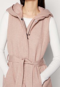 Světle růžová bezramenní vesta s kapucí, měkká textura, přední zip a stahovací šňůrka v pase. Pod ní je bílý top s dlouhým rukávem.