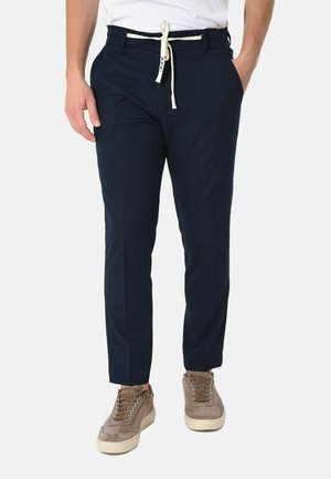 Uomo che indossa pantaloni blu navy con cintura bianca a coulisse e sneaker beige in camoscio, in piedi con una mano in tasca.