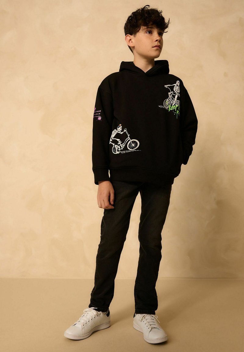 Garçon portant un sweat à capuche noir avec des squelettes à vélo imprimés dessus, un pantalon noir et des baskets blanches, se tenant devant un fond beige.