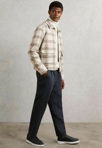 Ung mand iført en beige ternet jakke, hvid turtleneck, mørkegrå bukser og sorte sneakers, stående foran en ensfarvet lys grå baggrund.