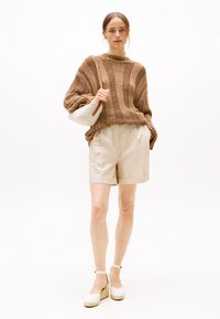 Bruin gebreide trui met brede mouwen, beige op maat gemaakte shorts en witte plateau sandalen. Houdt een kleine witte tas met een strap vast.