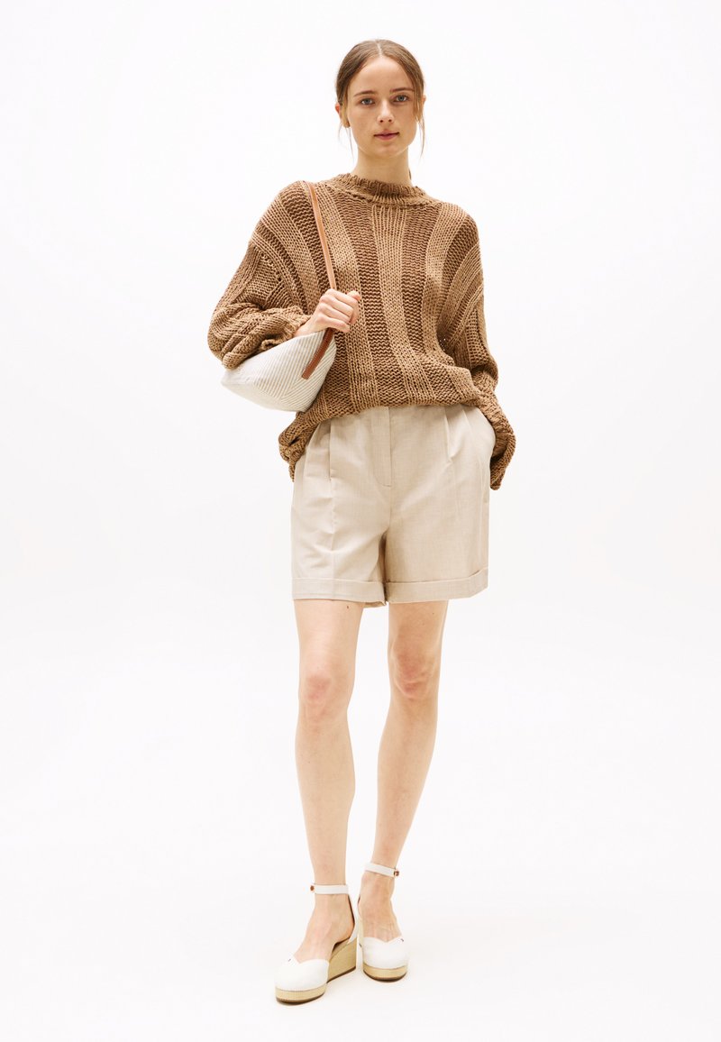 Bruin gebreide trui met brede mouwen, beige op maat gemaakte shorts en witte plateau sandalen. Houdt een kleine witte tas met een strap vast.