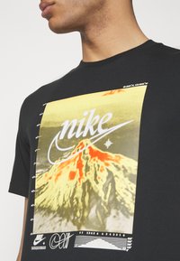 Svart t-shirt med en färgglad grafik av en vulkan med "Nike"-logotypen överlagd i vitt. Inkluderar koordinatdetaljer och designaccent.