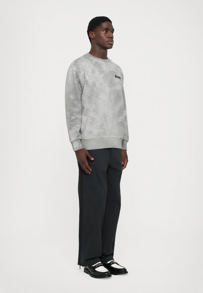 Mann steht da und trägt einen hellgrauen Batik-Pullover, lockere schwarze Hosen und schwarz-weiße Loafers mit goldener Kettendetails.
