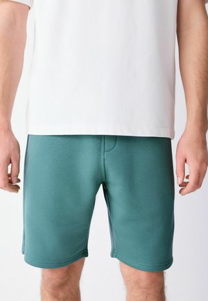 Mand iført petroleumsfarvede knælange shorts og en hvid kortærmet skjorte, stående med arme afslappet langs siden mod en ensfarvet baggrund.