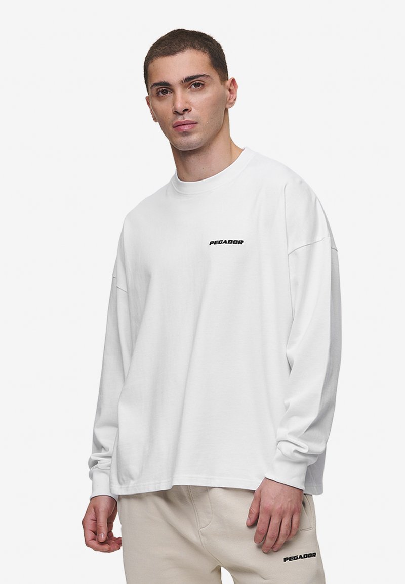 Camiseta blanca de manga larga hecha de algodón. Con un cuello redondo y un pequeño logo negro "PEGADOR" en el pecho. Corte casual con hombros caídos.