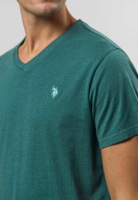 Teal v-ringad t-shirt tillverkad av mjukt, lätt tyg. Har en liten broderad logotyp på vänster bröst och korta ärmar.