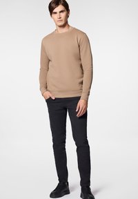 Beżowy sweter z okrągłym dekoltem noszony z czarnymi spodniami slim fit i czarnymi sneakersami. Tkanina wydaje się gładka, z prążkowanymi mankietami i dołem.