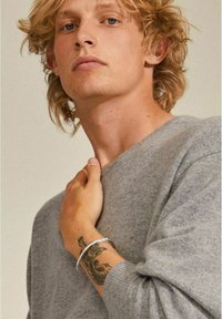 Maglione grigio realizzato in morbido tessuto, con scollatura a girocollo. Un bracciale d'argento sul polso mette in evidenza un tatuaggio dettagliato. Posizione rilassata e casual.