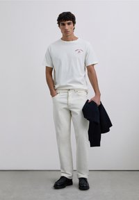 Una camiseta de algodón blanca con un diseño gráfico rojo, combinada con jeans crema y zapatos negros. El modelo sostiene un suéter oscuro. Fondo simple.