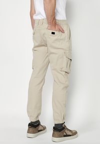 Beige Cargo-Hosen mit elastischen Bündchen, mehreren Seitentaschen und einer glatten Textur. Kombiniert mit braunen Sportschuhen und grauen Socken.