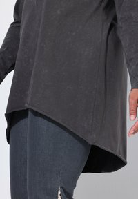 Dunkelgraues Langarmoberteil mit strukturiertem Finish, versehen mit abgerundetem Saum und Seitenschlitzen, kombiniert mit dunklen Jeans.