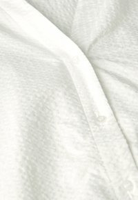 Camicia bianca con bottone, realizzata in un tessuto testurizzato con un motivo sottile. Presenta un collo a V e bottoni bianchi lungo la parte anteriore.