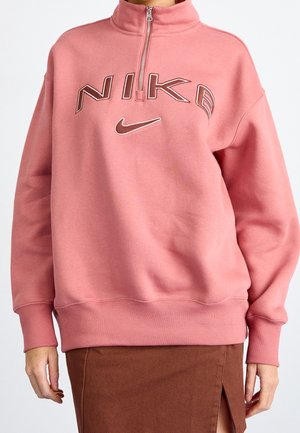 Personne portant un sweat Nike rose à fermeture éclair quart, avec une jupe marron fendue sur le devant, visible de la poitrine jusqu'à mi-cuisse.