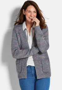 Femme souriante avec de longs cheveux bruns portant un cardigan en tricot multicolore sur une chemise blanche et un jean bleu, se tenant devant un fond blanc.