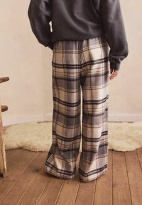Person wearing wide-leg beige, gray, and black plaid pants and a dark long-sleeve top standing on wooden floor near a chair and rug.