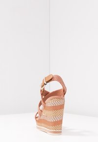 Sandale compensée tissée marron avec bride à boucle autour de la cheville et talon plateforme texturé, vue de l'arrière sur une surface blanche avec un fond blanc.