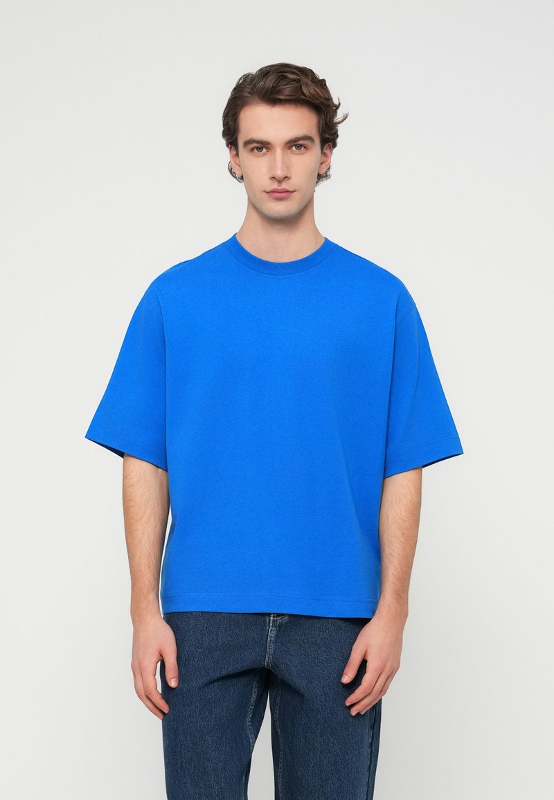 Μπλε βαμβακερό μπλουζάκι με κοντά μανίκια, στρογγυλή λαιμόκοψη και άνετη εφαρμογή. Χωρίς ορατά μοτίβα ή διακοσμητικά, σε συνδυασμό με μπλε τζιν.