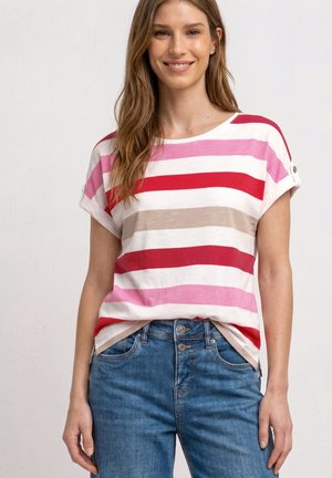 Uśmiechnięta kobieta w różowo-czerwono-beżowo-białym paskowanym krótkim topie wpuszczonym w niebieskie dżinsy na jednolitym tle.