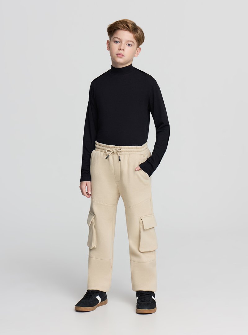 Czarna bluzka z długim rękawem, beżowe spodnie cargo z kieszeniami i ściągaczem w pasie oraz czarne trampki z białymi akcentami, stojące na szarym tle.