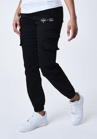 Pantaloni cargo neri con polsini elasticizzati, dotati di molteplici tasche laterali e ricamo bianco "Project x Paris" sul davanti. Indossati con sneaker bianche.